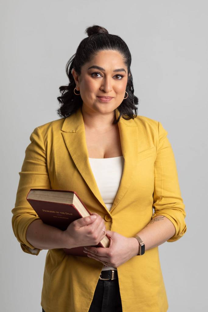 Profile photo of Jai, a South Asian woman with shoulder-length dark hair pulled back in a half-bun. She is wearing a yellow blazer and white blouse, medium hoop earrings, and is subtly smiling while looking at the camera. In her arms is a large 19th century book with the title "Problems of the Deaf" in gold print. 